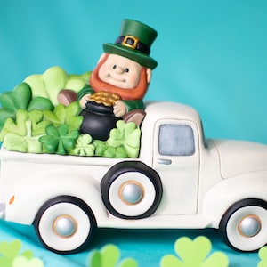 Op de afbeelding: Een witte keramische vrachtwagenfiguur met zwarte wielen, met een kabouter, een pot goud en groene klavers. De kabouter draagt een groene hoed en jas. De achtergrond is lichtblauw.