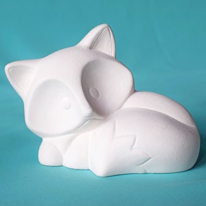 May include: White ceramic fox figurine with a curled tail. The fox is lying down with its head resting on its paws. The figurine is unpainted and ready to be decorated.