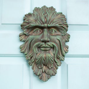 May include: A green and brown sculpted face with a leafy beard and hair, resembling a nature spirit or a green man. The face is mounted on a light blue door.