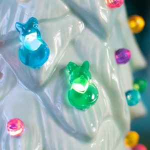 May include: A light blue ceramic Christmas tree with colorful lights and bunny ornaments. The tree is decorated with small, round, multi-colored lights.
