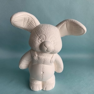 Puede incluir: Figurita de conejo de cerámica blanca con orejas largas y un diseño de pelaje texturizado. El conejo lleva un mono con un detalle cosido.