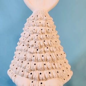 May include: A cream-colored ceramic Easter bunny-shaped decoration. The bunny has large upright ears and a textured, cone-shaped body with small holes. The base is a basket design. A decorative item for Easter.