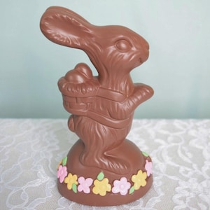 May include: A chocolate Easter bunny holding a basket of eggs, standing on a chocolate base decorated with pastel flowers.