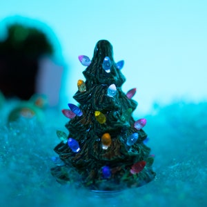 May include: A small green ceramic Christmas tree with colorful lights. The tree is sitting on a bed of white faux snow.