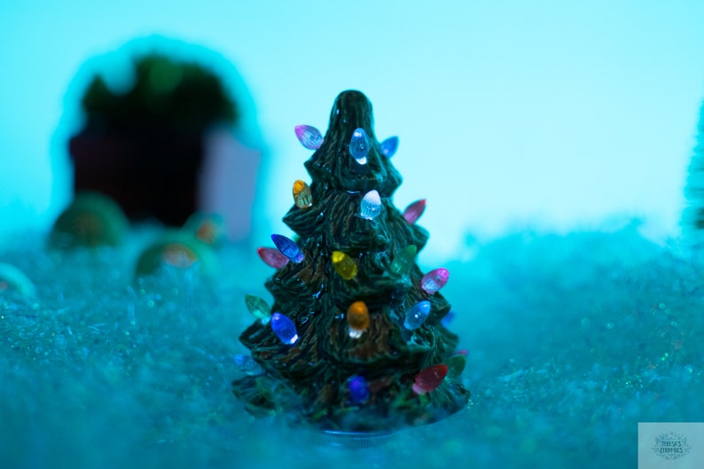 May include: A small green ceramic Christmas tree with colorful lights. The tree is sitting on a bed of white faux snow.