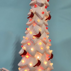 May include: A white ceramic Christmas tree with red cardinal ornaments and warm white lights. The tree is lit and has a cord for plugging in.