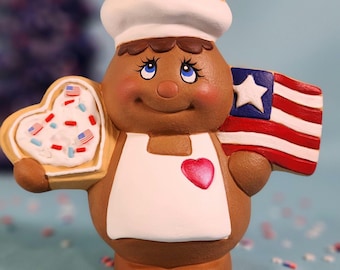 May include: A gingerbread person figurine wearing a white chef's hat and apron, holding a heart-shaped cookie with red, white, and blue sprinkles and a star-shaped cookie with red, white, and blue sprinkles. The figurine is also holding a small American flag.