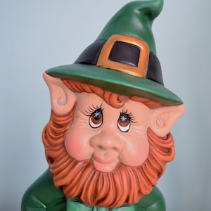May include: A green leprechaun figurine with a green hat, a brown beard, and a green bow tie. The figurine has a friendly expression and is wearing a green shirt.