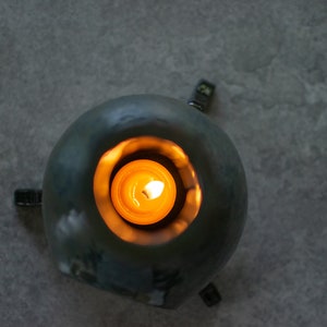 Pode incluir: Um porta-velas de cer&acirc;mica azul-esverdeado de forma redonda com quatro pequenos p&eacute;s de metal preto. Uma pequena vela redonda est&aacute; a arder dentro do porta-velas.