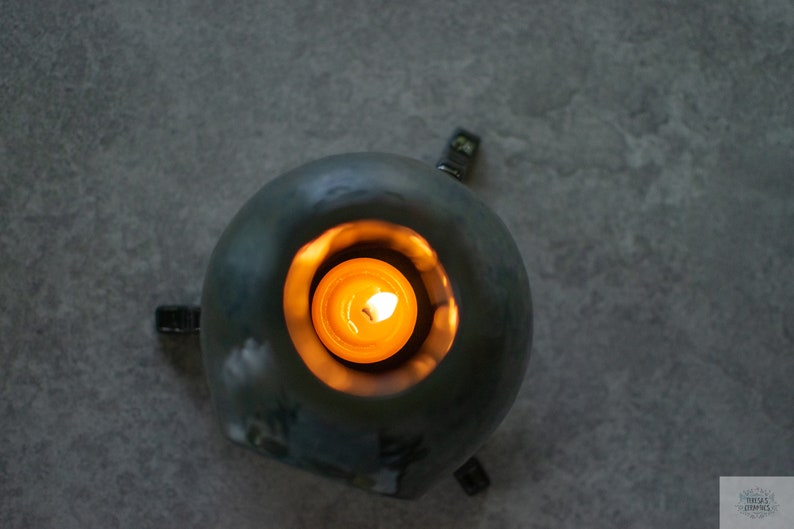 Pode incluir: Um porta-velas de cer&acirc;mica azul-esverdeado de forma redonda com quatro pequenos p&eacute;s de metal preto. Uma pequena vela redonda est&aacute; a arder dentro do porta-velas.