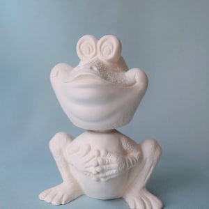 Pu&ograve; includere: Figurina di rana in ceramica bianca con occhi grandi e un ampio sorriso. La rana &egrave; seduta con le gambe incrociate e le mani giunte davanti a s&eacute;.