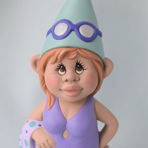 May include: A ceramic gnome figurine wearing a light blue hat with a purple visor and goggles. The gnome is wearing a purple swimsuit and has a light blue and purple patterned towel draped over one arm.