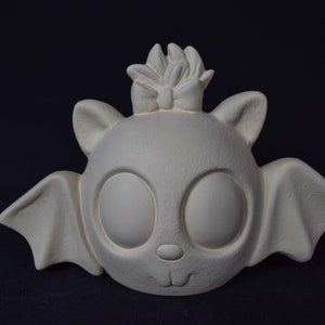 May include: White ceramic bat figurine with large eyes and a bow on its head. The bat has wings and a cute, smiling face.