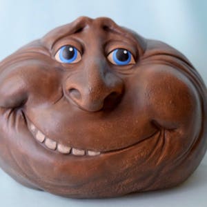 May include: A brown ceramic sculpture of a face with a wide smile and large blue eyes. The sculpture has a textured surface and a large, bulbous nose.