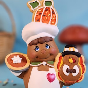 Pode incluir: Uma figurinha de pão de gengibre com um chapéu de cozinheiro com um biscoito de abóbora no topo, segurando uma torta e um biscoito de peru. A figurinha está cercada por confeitos em um fundo azul.