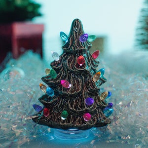 May include: A small ceramic Christmas tree with colorful lights. The tree is brown and has a textured surface. It is sitting on a bed of white and blue iridescent glitter.