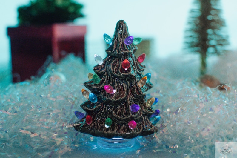 May include: A small ceramic Christmas tree with colorful lights. The tree is brown and has a textured surface. It is sitting on a bed of white and blue iridescent glitter.