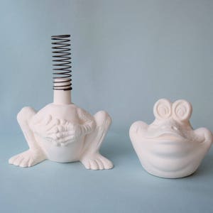 Pu&ograve; includere: Due statuine di rana in ceramica bianca. Una rana ha una molla a spirale sulla testa. L'altra rana ha una bocca larga e aperta.