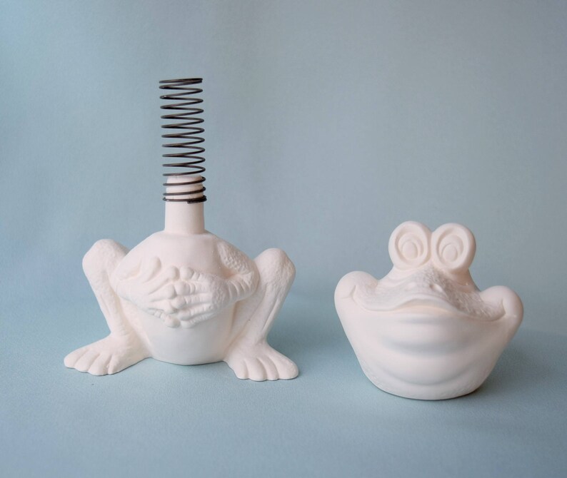 Pu&ograve; includere: Due statuine di rana in ceramica bianca. Una rana ha una molla a spirale sulla testa. L'altra rana ha una bocca larga e aperta.