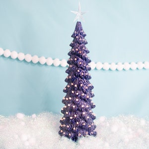 May include: A dark blue ceramic Christmas tree with white lights and a star on top. The tree is sitting on a white sparkly surface.
