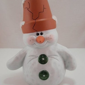 May include: A white ceramic snowman figurine with a brown flower pot for a hat. The snowman has a carrot nose, black eyes, and a smiling mouth. It has two green buttons for eyes.