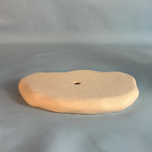 May include: A flat, irregularly shaped, light beige ceramic tray with a textured, matte surface. The object features a single, small, circular hole positioned slightly off-center on its top surface, with rounded, uneven edges throughout.