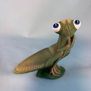 May include: A whimsical green figurine of an insect-like creature with large blue eyes. The figurine has a detailed wing and is posed on a green base. The color palette includes shades of green, brown, and blue.