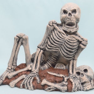 May include: A ceramic skeleton figurine with a skull resting on its lap. The skeleton is sitting on a brown dirt base with bones sticking out. The skeleton is painted in a light beige color.