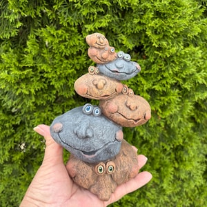 May include: A vertical stack of five sculpted stone-like figures with textured surfaces, each featuring distinct facial expressions, prominent noses, and small round eyes. The figures are colored in shades of gray and earthy brown, arranged in a tower.