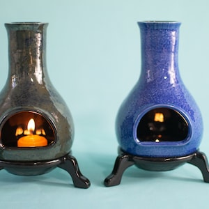 Pode incluir: Dois difusores de &oacute;leo de cer&acirc;mica com um esmalte azul esverdeado e azul. Cada difusor tem uma pequena abertura na frente para uma vela de ch&aacute;. Os difusores est&atilde;o em suportes pretos com tr&ecirc;s pernas.