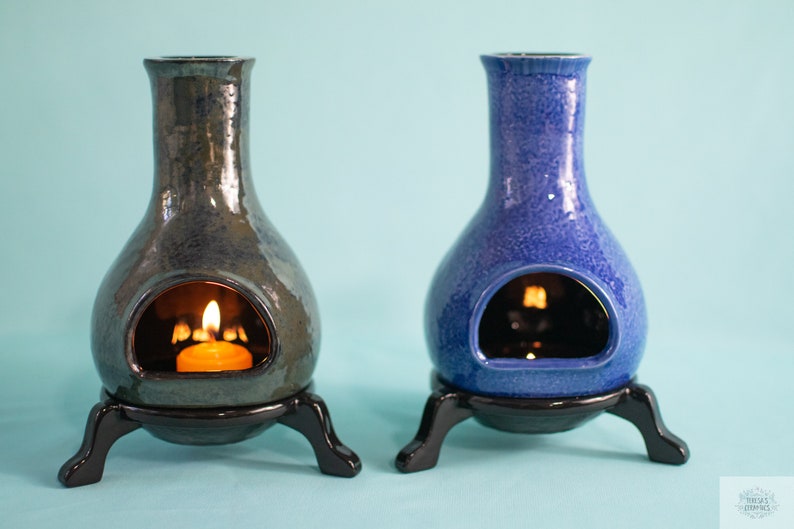Pode incluir: Dois difusores de &oacute;leo de cer&acirc;mica com um esmalte azul esverdeado e azul. Cada difusor tem uma pequena abertura na frente para uma vela de ch&aacute;. Os difusores est&atilde;o em suportes pretos com tr&ecirc;s pernas.