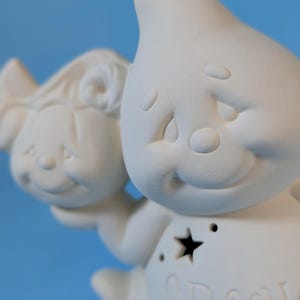 May include: A white ceramic figurine of a smiling ghost holding a smaller figure. The ghost has a star-shaped cutout and the word "Spooky" is written on the front. The background is a solid light blue.
