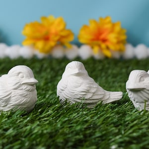 May include: Three white ceramic bird figurines on a green artificial grass surface. The birds are in various poses, with one sitting upright, one lying down, and one standing with its wings spread.