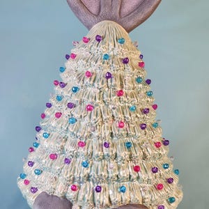 May include: A ceramic Christmas tree figurine with a bunny rabbit design. The tree is decorated with colorful lights and is sitting in a brown wicker basket.