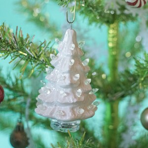 May include: A white ceramic Christmas tree ornament with a light-up feature. The ornament is hanging on a green artificial Christmas tree.