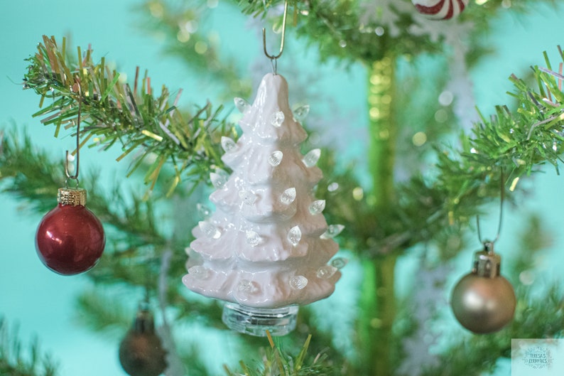 May include: A white ceramic Christmas tree ornament with a light-up feature. The ornament is hanging on a green artificial Christmas tree.