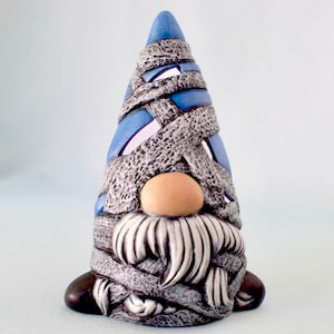 May include: A ceramic gnome figurine with a blue and gray hat and a white beard. The gnome is wrapped in bandages, giving it a spooky Halloween look.