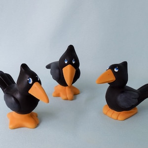 May include: Three black crow figurines with orange beaks and blue eyes. The crows are standing on a light blue surface.