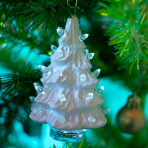 May include: A white ceramic Christmas tree with clear lights, hanging from a green Christmas tree branch.