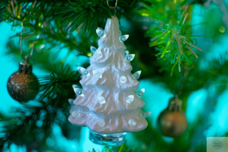 May include: A white ceramic Christmas tree with clear lights, hanging from a green Christmas tree branch.