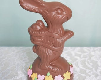 May include: A chocolate Easter bunny holding a basket of eggs, standing on a chocolate base decorated with colorful flowers.