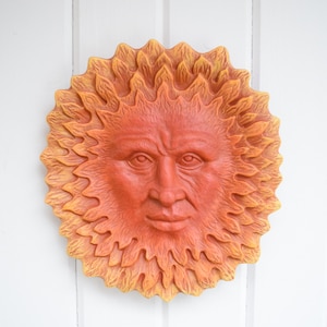 May include: A terracotta sun face wall decor with a detailed face and a yellow and orange sunburst design.