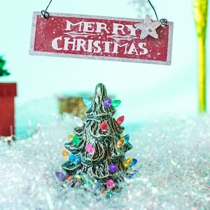 May include: A miniature Christmas tree with colorful lights sits on a bed of white iridescent tinsel. A red sign above reads "Merry Christmas" with a white star.