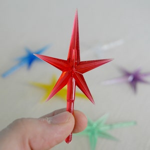 May include: A hand holding a red star-shaped ornament with a pointed tip. Other star ornaments in blue, yellow, purple, and green are out of focus in the background. The ornaments appear to be made of translucent plastic.