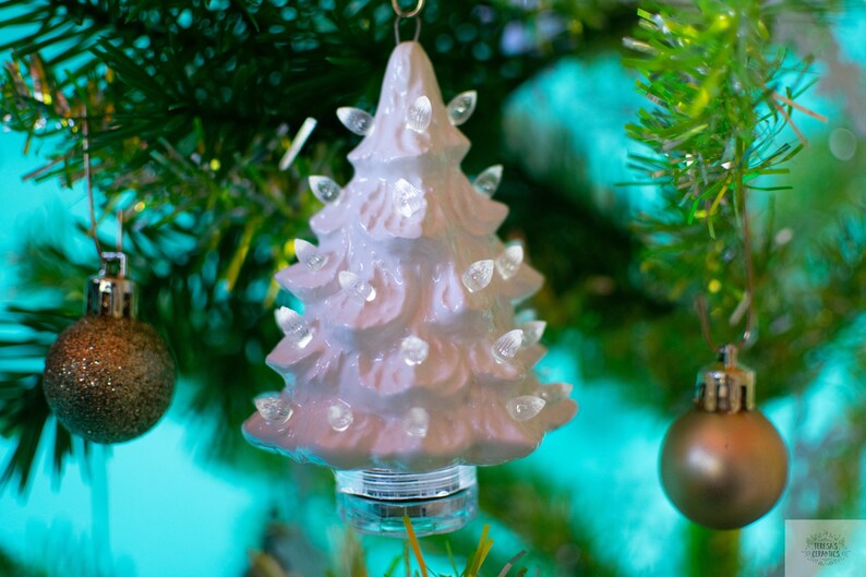May include: A white ceramic Christmas tree ornament with small white lights. The ornament is hanging on a green Christmas tree.