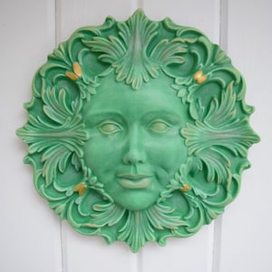 May include: A green ceramic wall plaque with a stylized face surrounded by leafy details. The face has a serene expression and gold accents.