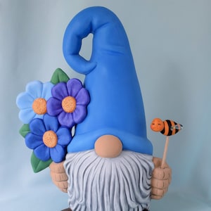 May include: A blue gnome figurine with a white beard, holding a bouquet of blue flowers and a bee on a stick.