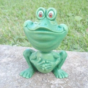 Pu&ograve; includere: Una statuetta di rana verde con un ampio sorriso e grandi occhi rotondi. La rana &egrave; seduta con le gambe incrociate e le mani giunte davanti a s&eacute;.