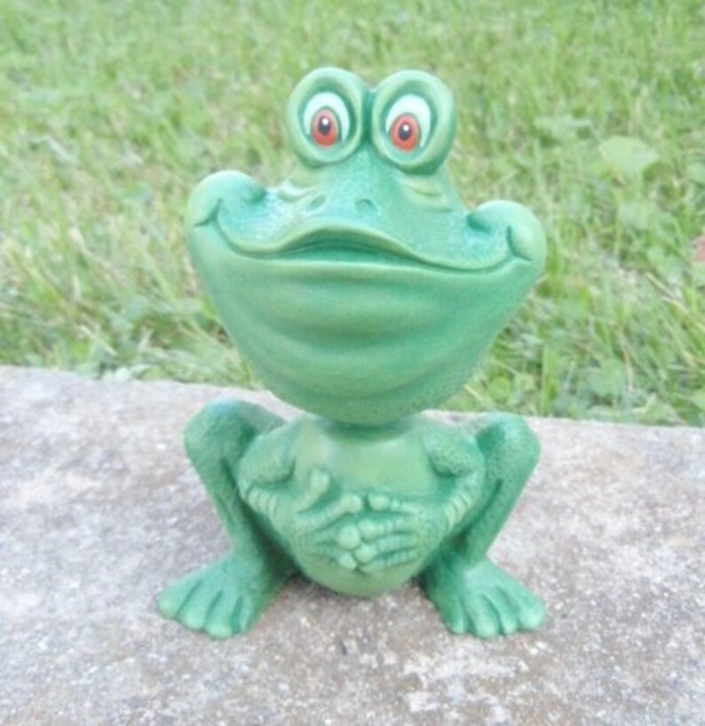 Pu&ograve; includere: Una statuetta di rana verde con un ampio sorriso e grandi occhi rotondi. La rana &egrave; seduta con le gambe incrociate e le mani giunte davanti a s&eacute;.