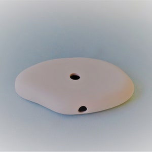 May include: A light pink ceramic dish with a rounded, irregular shape. The dish has two small holes, one in the center and one on the side.